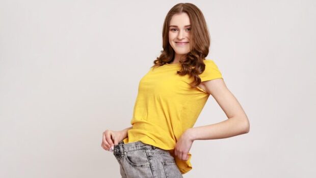 side-view-portrait-young-woman-big-jeans-yellow-casual-tshirt-showing-successful-weight-loss-her-slim-waist-looking-smiling-camera-indoor-studio-shot-isolated-gray-background_416530-23303